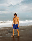 Man wearing blue swim shorts standing on a beach with waves in the background