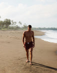 Man walking on a sandy beach with ocean and trees in the background. He is wearing Mens sustainable Swim shorts in dark red