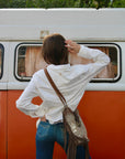 A woman viewed from behind stands in front of an orange and white vintage van, wearing a white button-up shirt, blue jeans, and carrying a brown fringe shoulder bag with a patterned strap.