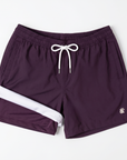 Flat lay image on a white backdrop of Men's purple swim shorts. The left leg of the swim shorts if rolled up to show the built in boxer lining.