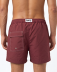 Burgundy swim shorts with a visible brand logo in the centre of the back of the waistband. A back pocket is visible, designed with subtle white stitching for a luxury feeling. The image is shot on a white background.