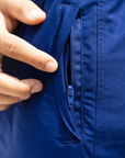 Men's swim shorts in blue. A close up image is showing a man's hand holding a zip to show the that the swim shorts have secure pockets for goods. Image photographed in a studio
