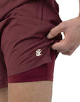 Burgundy men's swim shorts with a logo on a white background. The shorts are being lifted up slightly to show the anti-chafe swimwear lining.