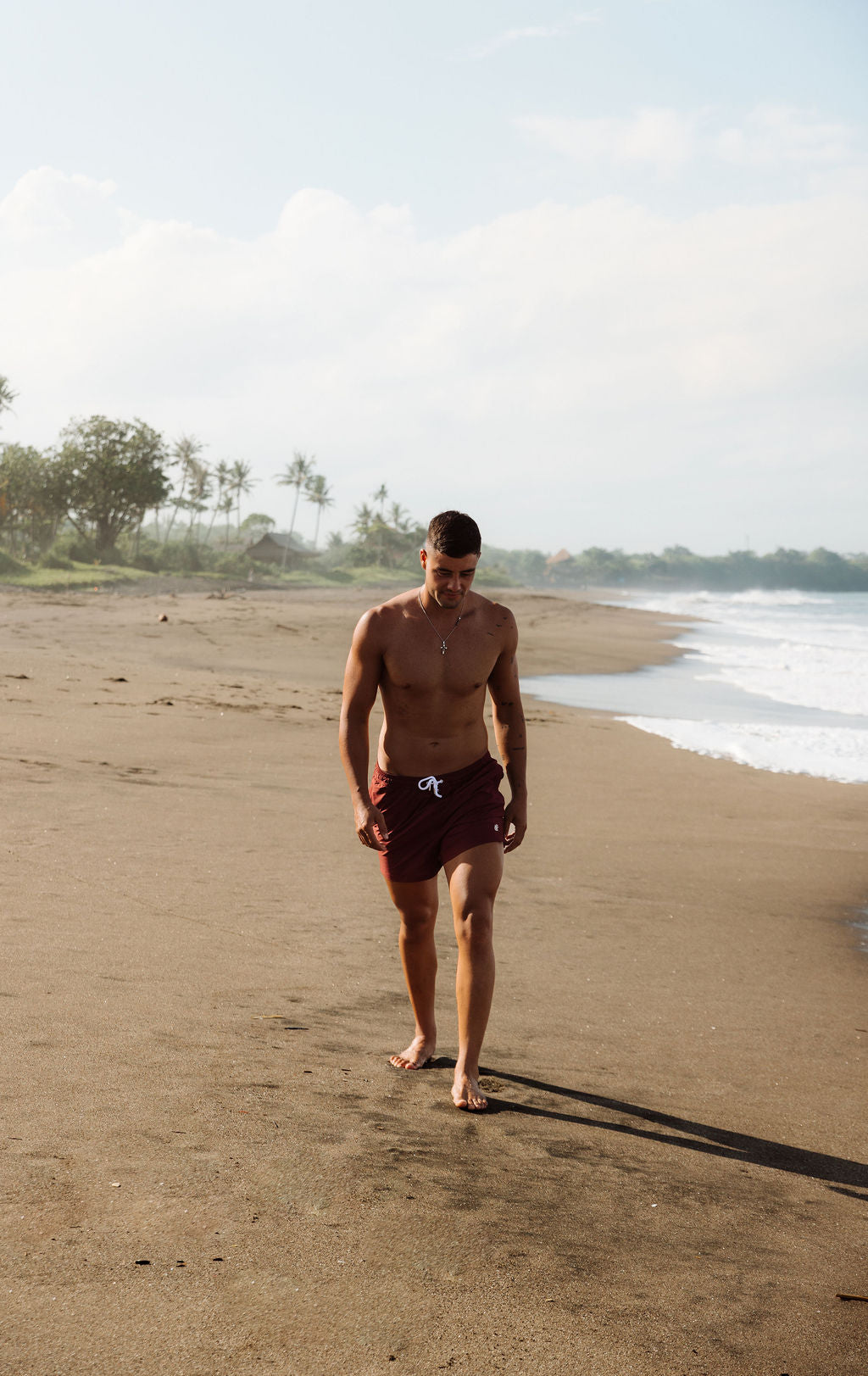 Man walking on a sandy beach with ocean and trees in the background. He is wearing Mens sustainable Swim shorts in dark red