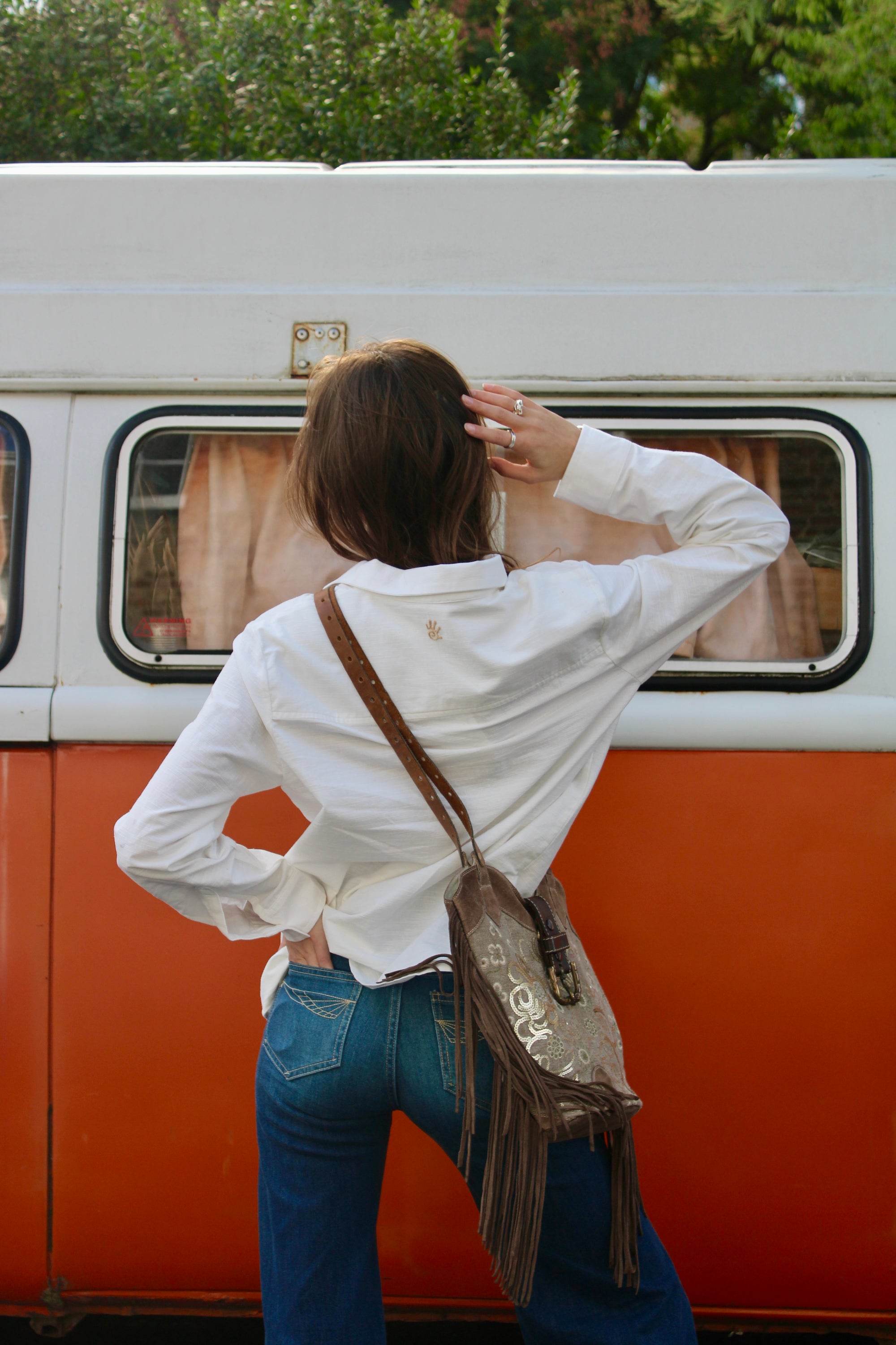 A woman viewed from behind stands in front of an orange and white vintage van, wearing a white button-up shirt, blue jeans, and carrying a brown fringe shoulder bag with a patterned strap.