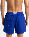 Blue swim shorts with a visible brand logo in the centre of the back of the waistband. A back pocket is visible, designed with subtle white stitching for a luxury feeling. The image is shot on a white background.
