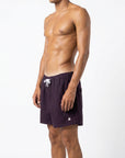 Model stands side on, showing the men's purple swimshorts from a side angle. The white Raw Potential emblem is shown in the image, along with the white draw string and adjustable waistband 